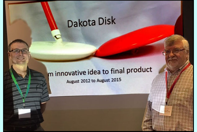 Paul and Gord are standing in front of their powerpoint which shows a red disk and a white disk each about 10 inches across and 1-2 inches high.  The white disk is attached to a cane and the slide says'Dakota Disk - From innovative idea to final market, August 2012 to August 2015'.